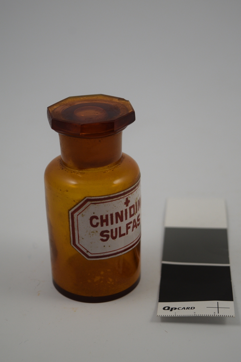 Brun glasskrukke med glasspropp. Etikett hvit, med rød skrift. Ett kors, betyr at innholdet er giftig. Chinidini Sulfas ble brukt i hjertemedisin.