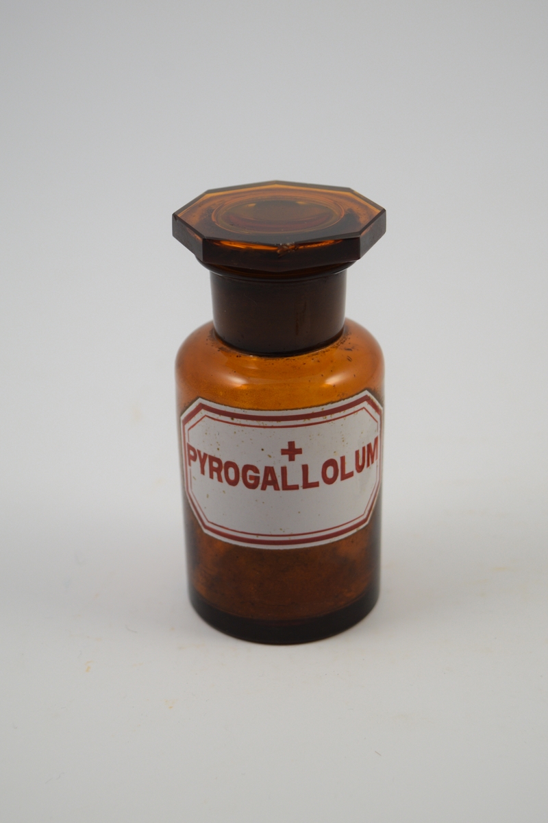 Glassflaske, brun med brun propp. Påført etikett; hvit med rød skrift. Ett rødt kors - betyr at innholdet er giftig. Pyrogallolum, "tretjære", ble brukt i salver mot eksem.