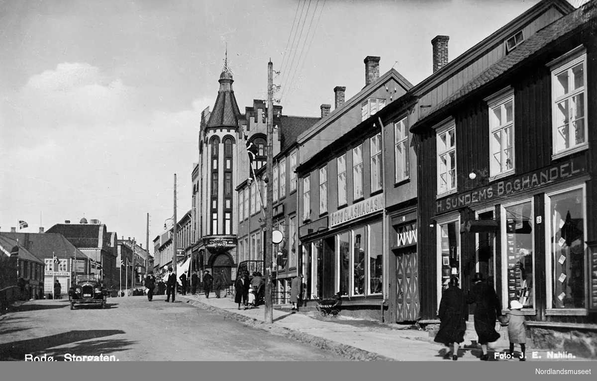 Postkort. Bodø, Storgata med H. Sundems bokhandel nærmest til høyre, Glassmagasinet og Tandberggården. O. Johanson, Tjærandsens jernvareforretning. På den andre siden av gaten ses Gustav Moe's jernvareforretning.
