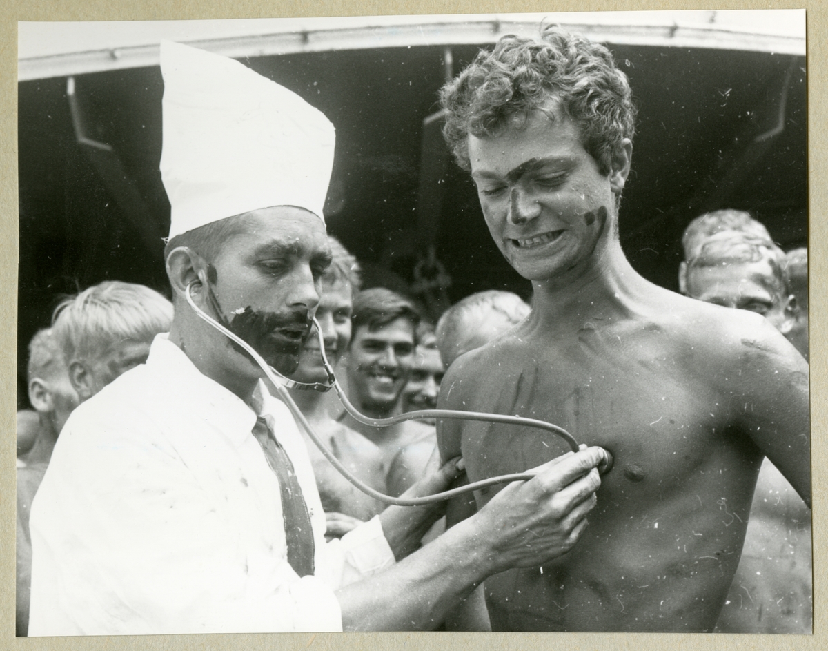 Bilden föreställer dåvarande kronprins Carl Gustav, den senare kung Carl XVI Gustaf, som blir undersökt av en man utklädd till läkare under ceremonierna kring ett linjedop ombord på minfartyget Älvsnabben. I bakgrunden syns barbröstade åskådare från besättningen. Bilden är tagen i samband med långresan 1966-1967.