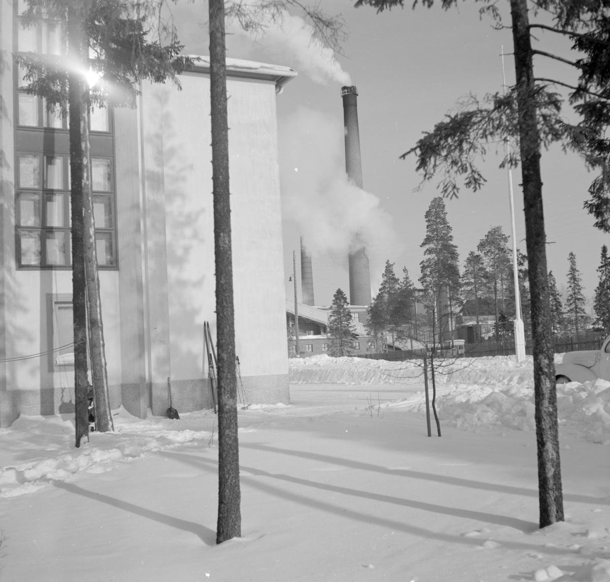 Exteriör vid F 19, Svenska frivilligkåren i Finland. Del av byggnad och träd. I bakgrunden syns Kemi pappersmassefabrik.