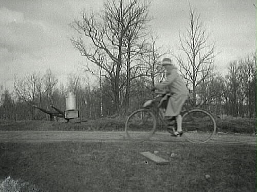Dam som cyklar på landsväg och ett mjölkbord med mjölkkanna i bakgrunden. 1920-tal.