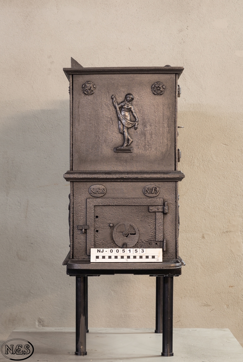 Kokeovn i to etasjer med eget fyrkammer og kokerom. Kokerommet kan åpnes med to dører på langsiden og med en luke i topplaten. Ovnsdøren til fyrkammeret er på kortsiden 1 etg. På langsidene i 1. etg er det tre muser og en engel med lette draperier på en enkel profillist. På kortsidene 2. etg er det draperikledd dansende muse på enkel profillist med en rosett i hvert øvre hjørne. Langsidene i 2.etg er dekorert med en rosett i hvert øvre hjørne.