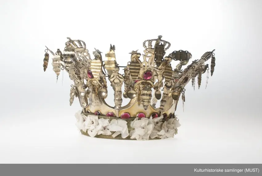 Brudekrone av sølv, forgylt. Nederst en glatt, innvendig stoppet ring, over
kronens 7 takker som munner ut i  Christian VII kronede monogram holdt av 2 løver,
og hver prydet med 4 røde glasstene og 1 mannshode med turbuan som holder et riflet
"løv". Til monogrammene er dessuten festet 3 "løv" og 2 skåler. Mellom
takkene en fløytespillende herme og en hane holdende et "løv". All
dekorasjon er presset og hvert takkestykke er arbeidet for seg og hengslet
til de øvrige. Det hele er avstivet med en messingtråd trukket gjennom
fastloddede øier på baksiden. Stemplet LH = Lauritz Hammer.1862.