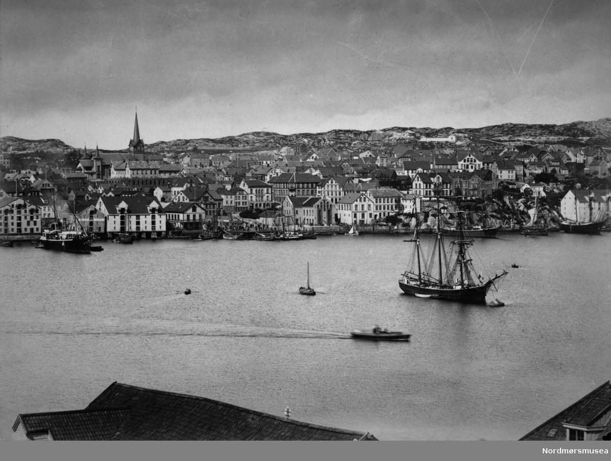 Fartøyet som ligger oppankret i forkant er en skonnertbark. Skipet til venstre har skorstein fra Bergenske Dampskipselskap.  Kirka, Piren, Blixhammeren. Kristiansund ca 1900. Nordmøre museums fotosamling.
