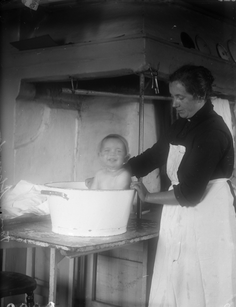 Tora Alinder badar Tore Alinder i balja på bord i köket, Sävasta, Altuna socken, Uppland 1925