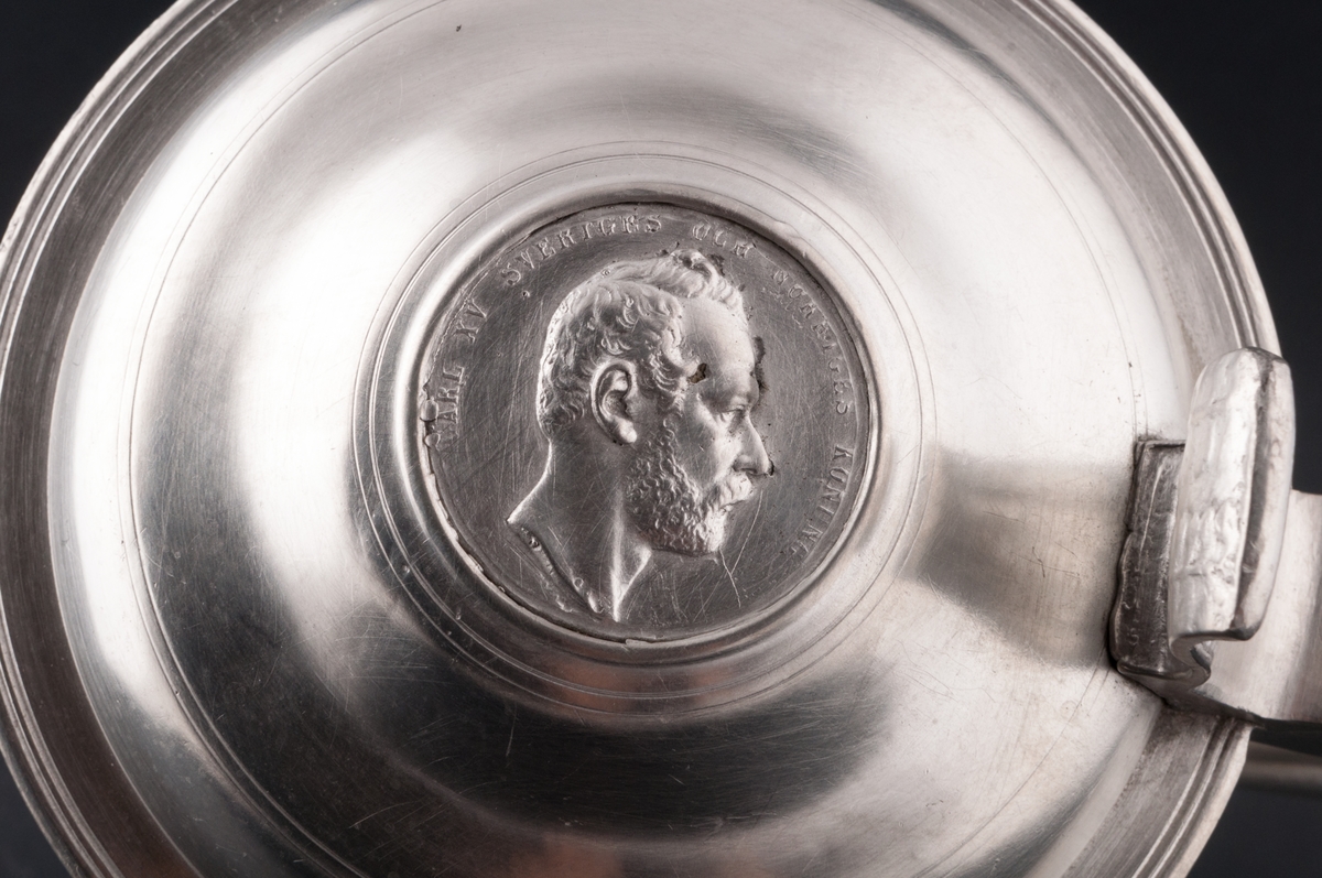 Bordkanna, tenn.
Ornerad med "DRICK BROR" på sidan. På locket mynt med Karl XV Sveriges och Norges konung. Stämplad i botten med "IS" (1863).