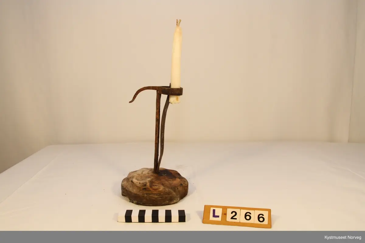Lysestake med rund, utskåret fot av tre. Lysholderen er smidd av jern, og har en klypemekanisme for å holde fast talglyset