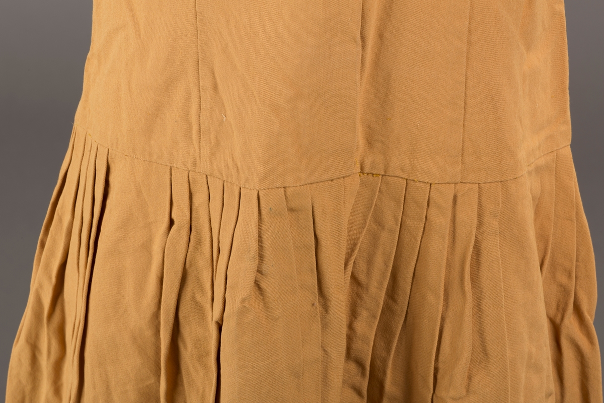 Form: Korte ermer, livet rynket til bærestykke foran, plissert skjørt, tre pynteknapper, en trykknapp, forlomme.