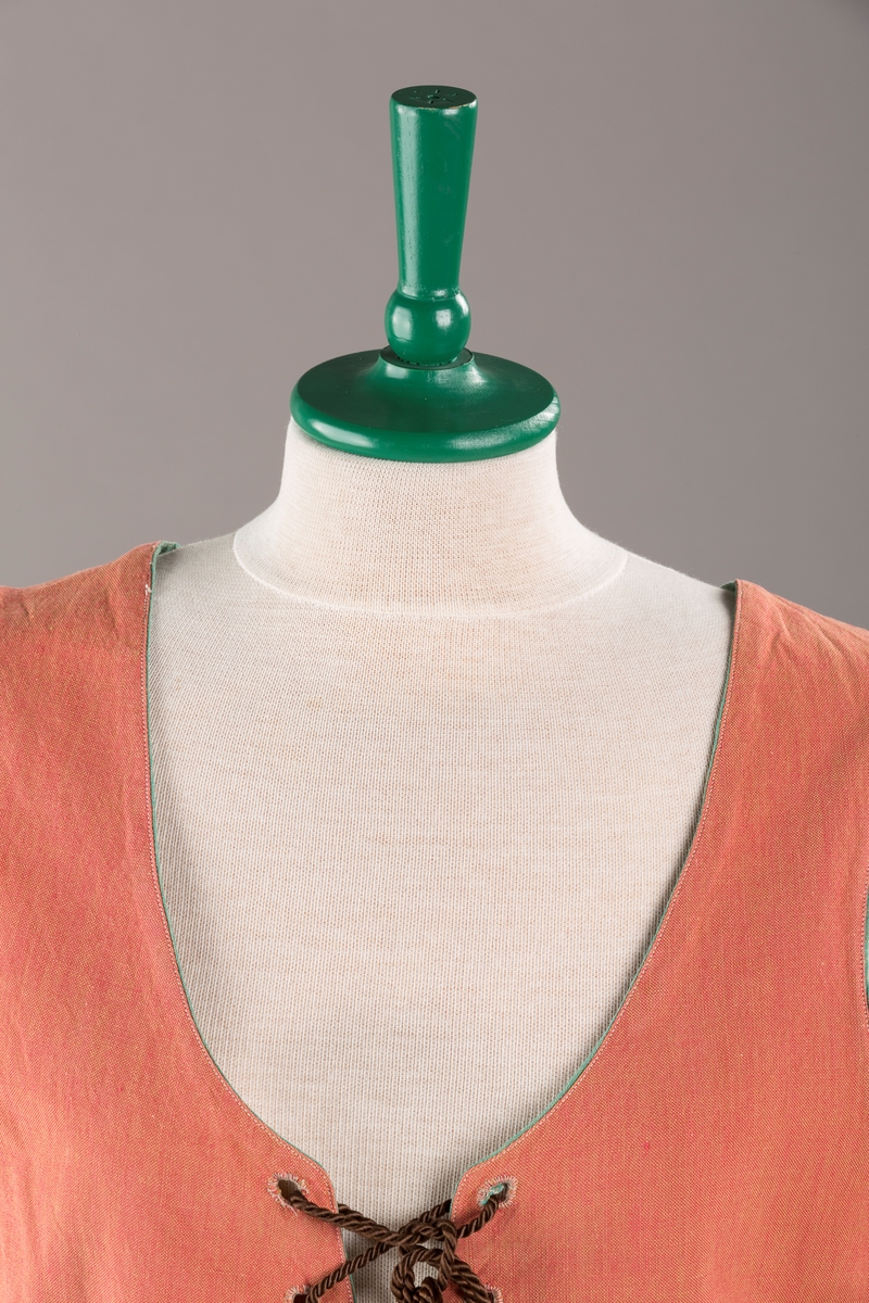 Rustrødt liv med grønn kant og fór, seks skjøter. Håndsydd forsterkning i kontrastfarge øverst i skjøtesplittene. Lukkes med tinnhekte og silkesnor.