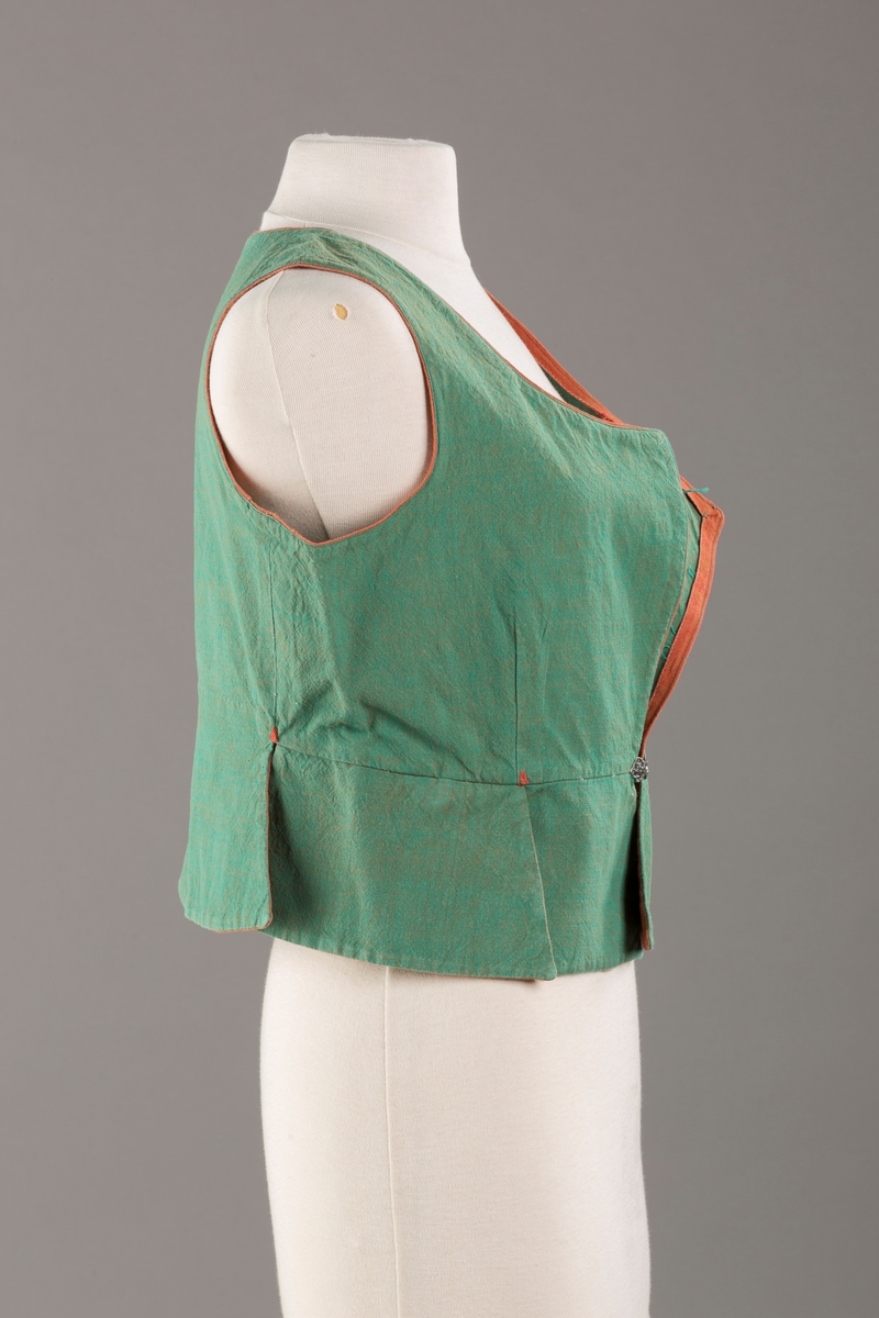 Grønt liv med rustrød kant og fór, seks skjøter. Håndsydd forsterkning i kontrastfarge øverst i skjøtesplittene. Lukkes med tinnhekte.