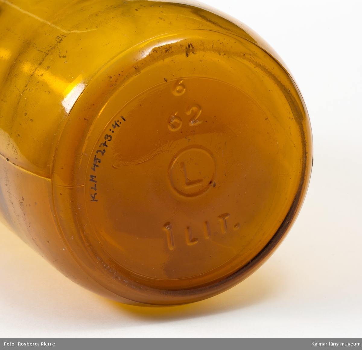 KLM 45273:4:1 Flaska för filmjölk, av glas, brunt. Volym 1 liter. Botten präglad med, 6 62 L (inom en cirkel) 1 LIT. Blå aluminiumkapsyl präglad med texten, Kalmarbygdens mejeriförening 23 Filmjölk.