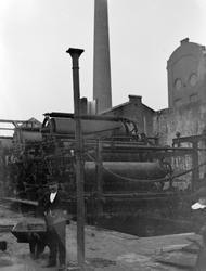 Mann med trillebår utenfor en fabrikk med høy pipe.