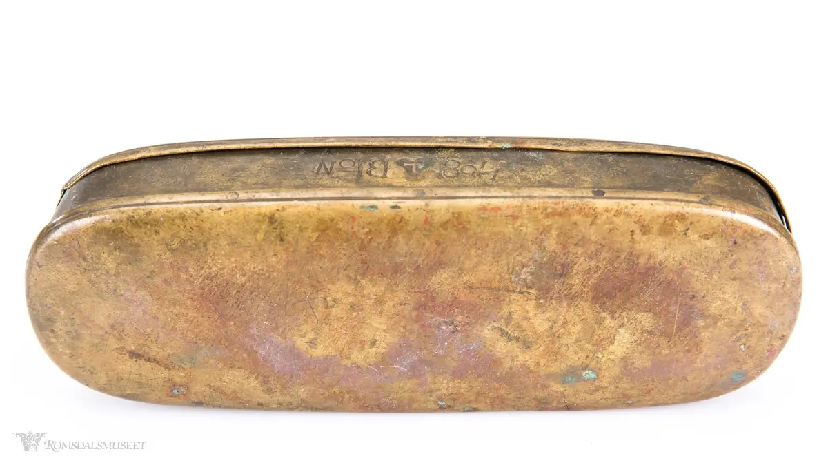 Rektangulær messingeske med avrundede ender. Har en påsatt og påklinket utenpåliggende bunn og utenpåliggende, hengslet lokk