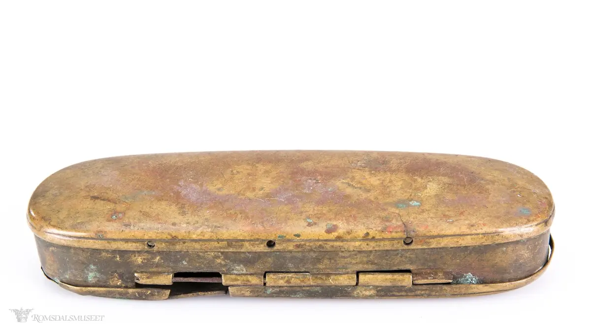 Rektangulær messingeske med avrundede ender. Har en påsatt og påklinket utenpåliggende bunn og utenpåliggende, hengslet lokk
