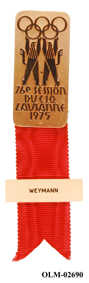Todelt badge fra den 76. IOC-Session i Lausanne 1975. Den øverste metalldelen har de olympiske ringer som holdes oppe av to bjørner med tekst under. Det er festet et rødt bånd under med navnemerke.
