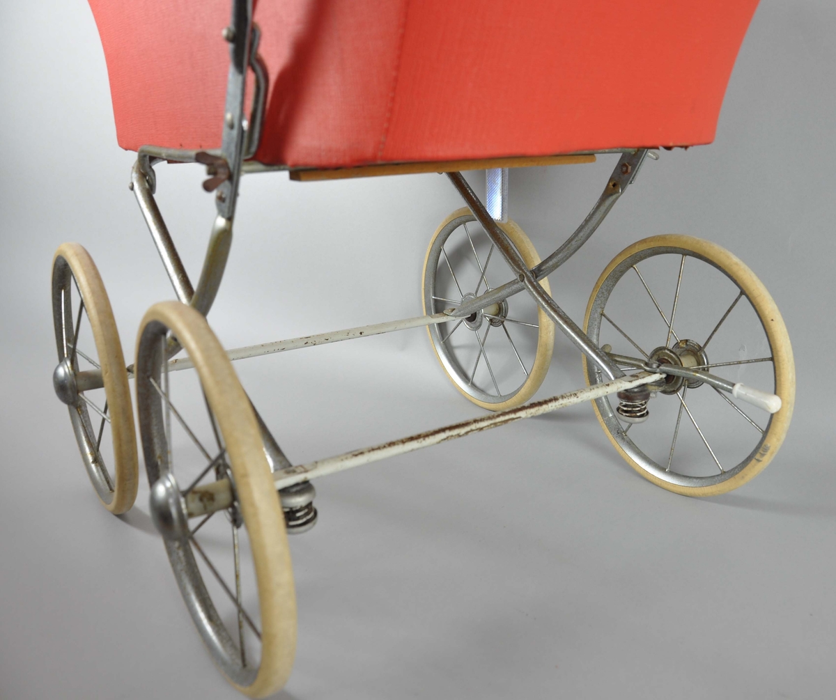 Dukkevogn med hjemmelaget madrass, dyne og pute. Vognen er av rød polyester med sammenleggbar kalesje. På sidene er det håndtak til å feste et løst vogndekke. Håndtak av kromt metall går ned på sidene av vognen, øverste del er lagt med hvit plast. Hjulene er hvite med eiker, ved høyre hjul er det en bremsespenne. Understellet på vognen er en lys treplate. På høyre håndtak er det satt på en refleks, av nyere tid.