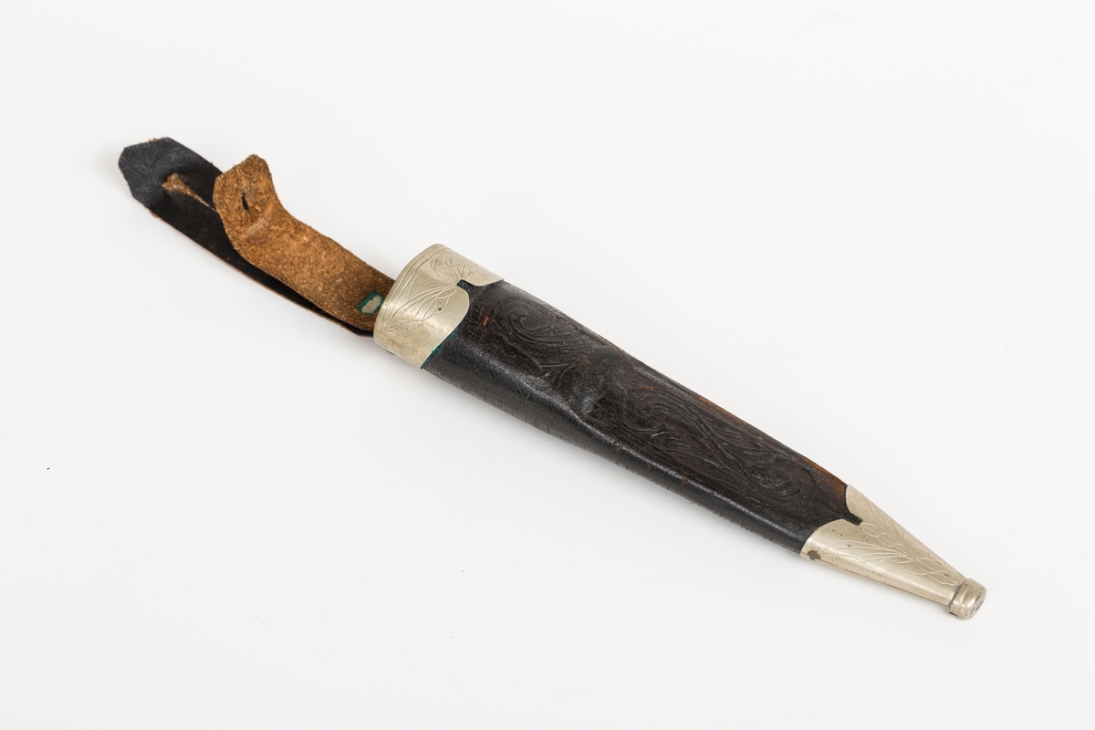 Håndsydd lærslire med holker oppe og nede. Slire og stropp av separate lærstykker. Sliren har enkel  pautet dekor. Holkene er håndloddet nysølv (sansynligvis) med enkel trambulert dekor. Toppholken har påloddet hengsle og D-ring til feste for slirestropp.