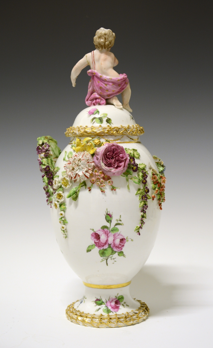 Potpourrikrukke av porselen med stett og lokk. Lokket er dekorert med håndmalt blomsterdekor og langs kanten en bord med blader i tre høyder dekorert med gull, over bladene gullfargede små kuler. På toppen av lokket en kjerub eller kupid som sitter på sitt ene kne. Han har på seg et rosa lendeklede og har holdt i et skjold eller en atributt. 
Urnens korpus er dekorert med håndmalte blomster, og modellerte blomster som sitter i klynger og strekker seg nedover korpus. De modellerte blomstene er uglasert og håndmalt. Urnen mangler en hanke. Den eksisterende hanken har modellerte blomster ved festene som strekker seg nedover i klynger.  Langs stettens kant samme bladbord som på lokket.