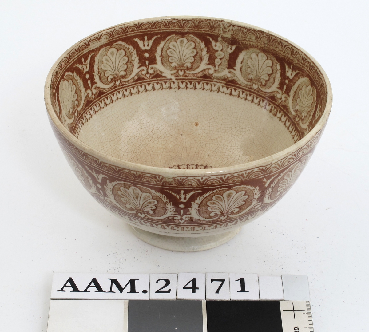 Bolle, stentøy, Buede sider, innsvunget fotring; trykt mønster i  brunt: Innv. og utv. bred ornamental bord m. skjell og "Garrya Eliptica" i bunnen lignende dekor, her er skjellene plassert i korsform i et rundt felt. Tilst. juli 1961: Krakellert.