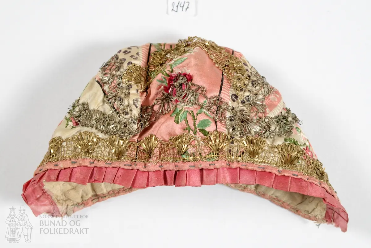 Dåpslue. Av rosa og hvit silkedamask med brosjert blomstermønster i to ulike rosa nyanser og grønt. Dekorert med metallkniplinger, i både sølv og gull. Smal rosa silkekanting rundt hele lua, i tillegg er et mørkerosa silkebånd i satengbinding foldelagt langs framkanten.  Foret med naturfarget linlerret. For og lue er vrangsydd, og så sydd sammen. Sømmene fra kniplingene er synlige på vrangen. Kun håndsøm. Stoffet er utnyttet med flere små skjøter.