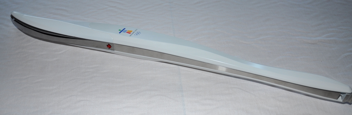 Hvit håndfakkel av plast og metall. Fakkelen har emblemet for de olympiske vinterleker i Vancouver i 2010. Det er også motiv av lønneblad på fakkelen.