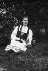 Portrett. Ung kvinne i folkedrakt sitter på gressbakken. Kar