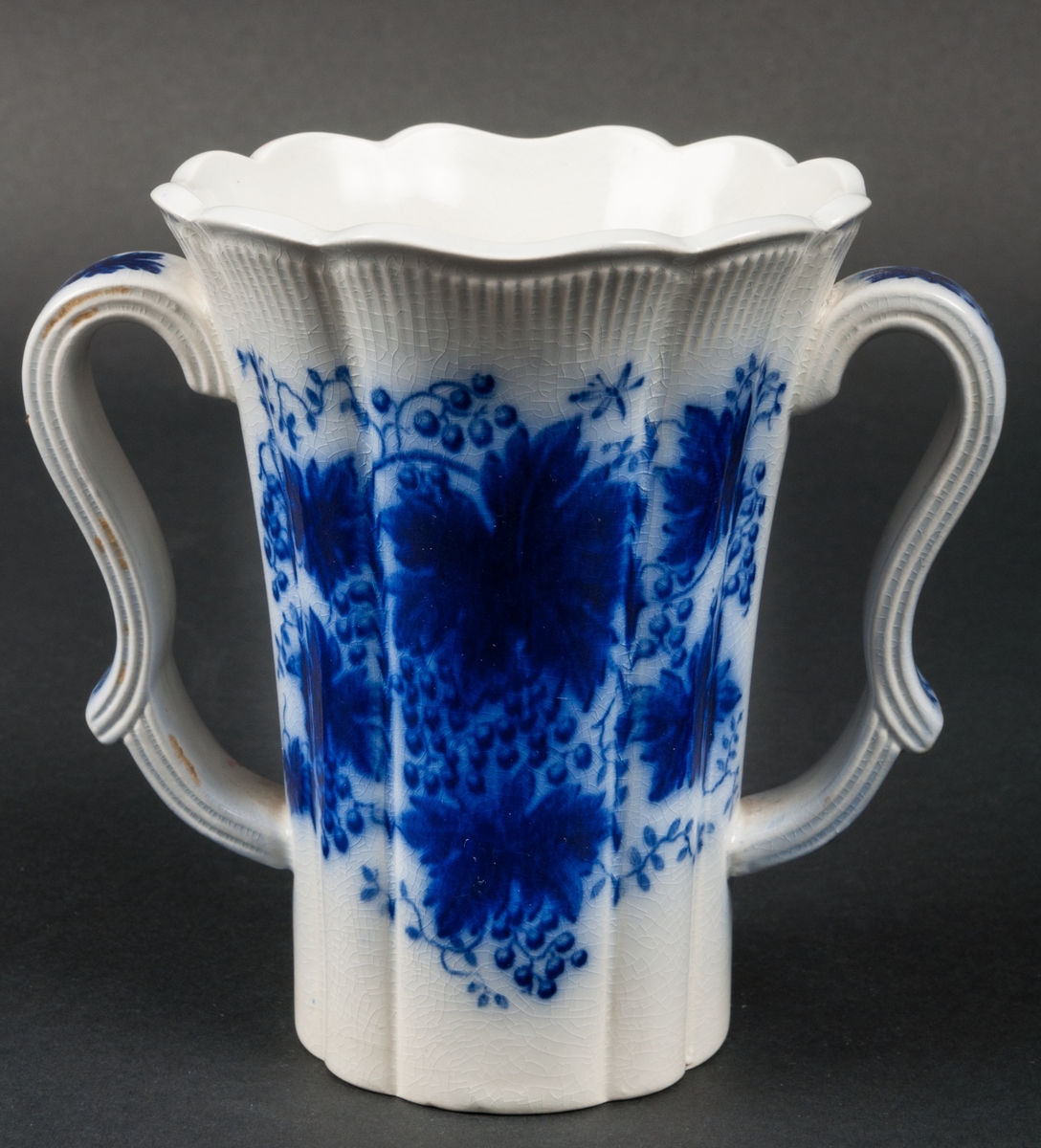 Vas, formgiven av Arthur Percy för Gefle Porslinsfabrik. Trattformad med vågig kant och buk. Två profilerade handtag. Dekor: Vinranka, blå.