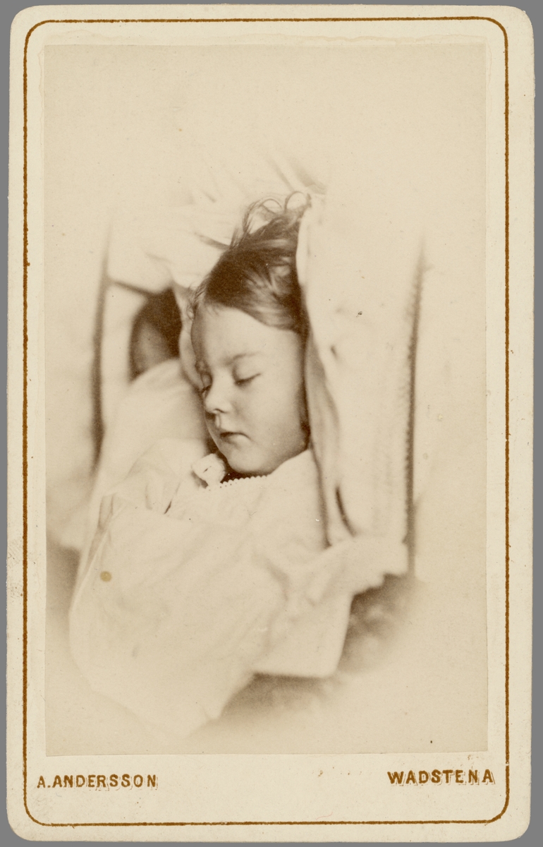 Post mortem-porträtt av Esther Wallin. Dotter till lasarettsläkaren i Vadstena, Axel Wallin och hans hustru Amalia Wallin. Flickan var parets förstfödda barn, men lyckan bestod endast i 1 år, 10 månader och 26 dagar. Tyfusen släckte hennes liv den 17 februari 1874.