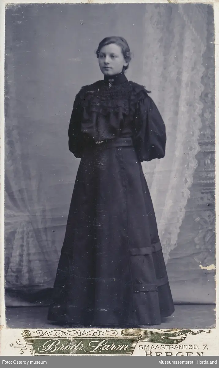 atelierfotografi av ung kvinne i heilfigur,  kledd i sid svart kjole (skjørt-bluse?) med håret opprulla, kapper og nål i halsen