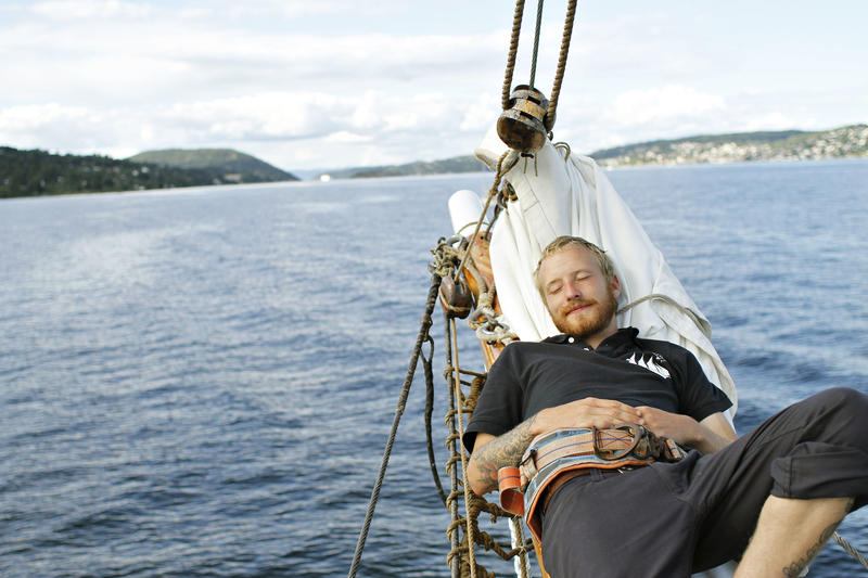 Mann slapper av ombord skonnerten "Svanen", blått vann, land og klar himmel i bakgrunnen.