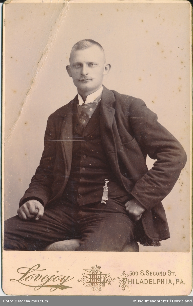 atelierfotografi av mann med bart i halvfigur som sit på ein krakk, kledd i jakke, vest, bukse, kvit skjorte og blomstra halstørkle