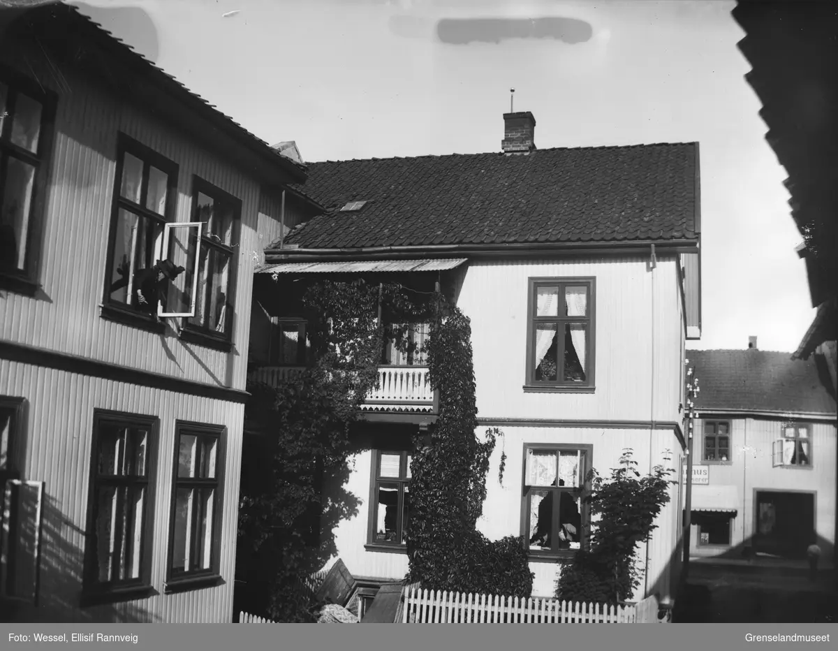 Stort byhus, slyngplanter på veggene, en kvinne henger ut av vinduet i andre etasje og en kvinne står nede på bakken ved en åpen kjellernedgang.