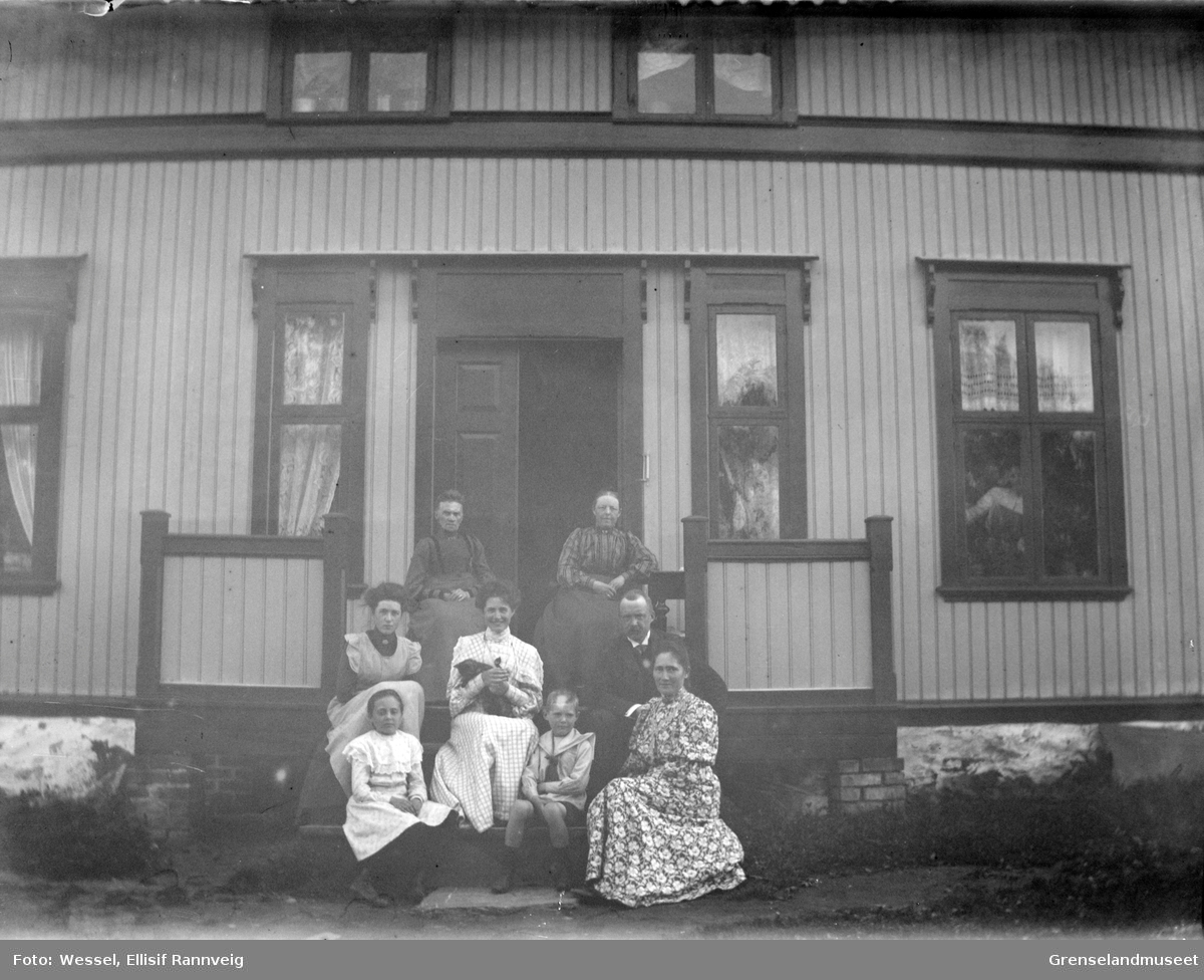 En gruppe mennesker fotografert sittende på en trapp utenfor et stort hus. To menn, fire kvinner og to barn. En kvinne ses i vinduet. Ukjent sted.