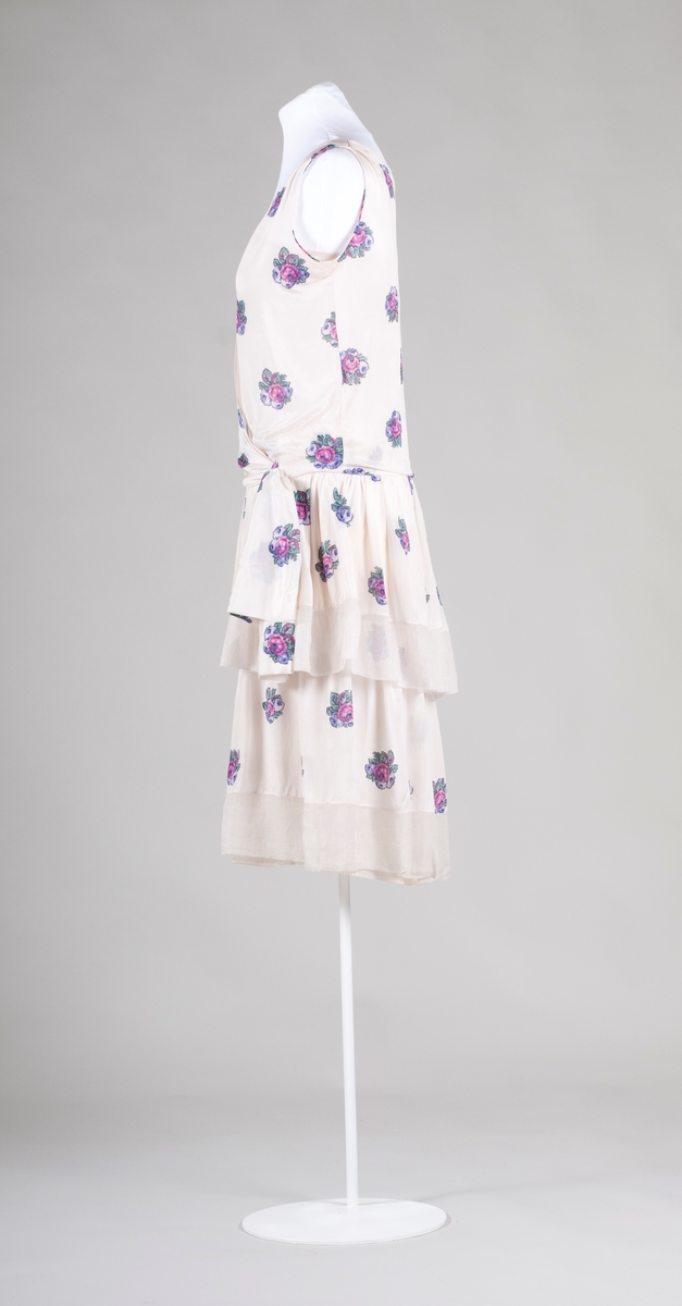 Klänning av vit siden med blomtryck. Kjol med volang och underklänning av tyll. Framstycket går omlott och avslutas med rosett i sidan.  V-formad halslinning. Ärmlös. 1920-tal.