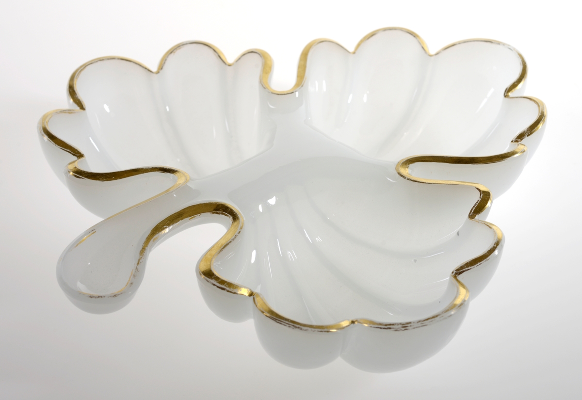 Skål av hvitt glass (opalinglass) formet som et eikeblad, med tre deler som former hver sin adskilte skål. Gullmalte kanter.