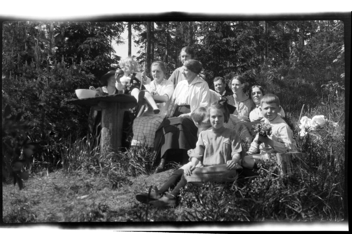 Alfhild Ferslew, Rolf Sundt jr., Hilda Sundt, Birgit Aubert, Birgitte Aubert, Bernt Ferslew, Anna Ferslew, Julie (Lillemor) Thaulow Aubert og Caspar Ferslew sitter sammen med ukjente mennesker på piknik i skogen. Fotografert 1922.