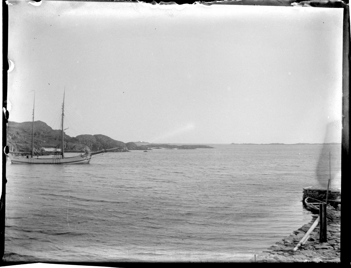 En seilskute ligger ankret opp innenfor Sandøy, og en robåt ligger ved en brygge i Loshavn; i bakgrunnen sees Einarsneset, Farsund. Fotografert 1900.