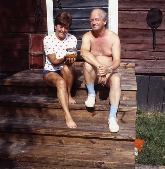 Tidningen LAND gjorde ett reportage i Hudiksvallstrakten, 29 juli 1986. Här ser vi en man och en kvinna som sitter i solen på en trätrapp till ett timrat hus. Kvinnan håller fram en konserveringsburk med innehåll.