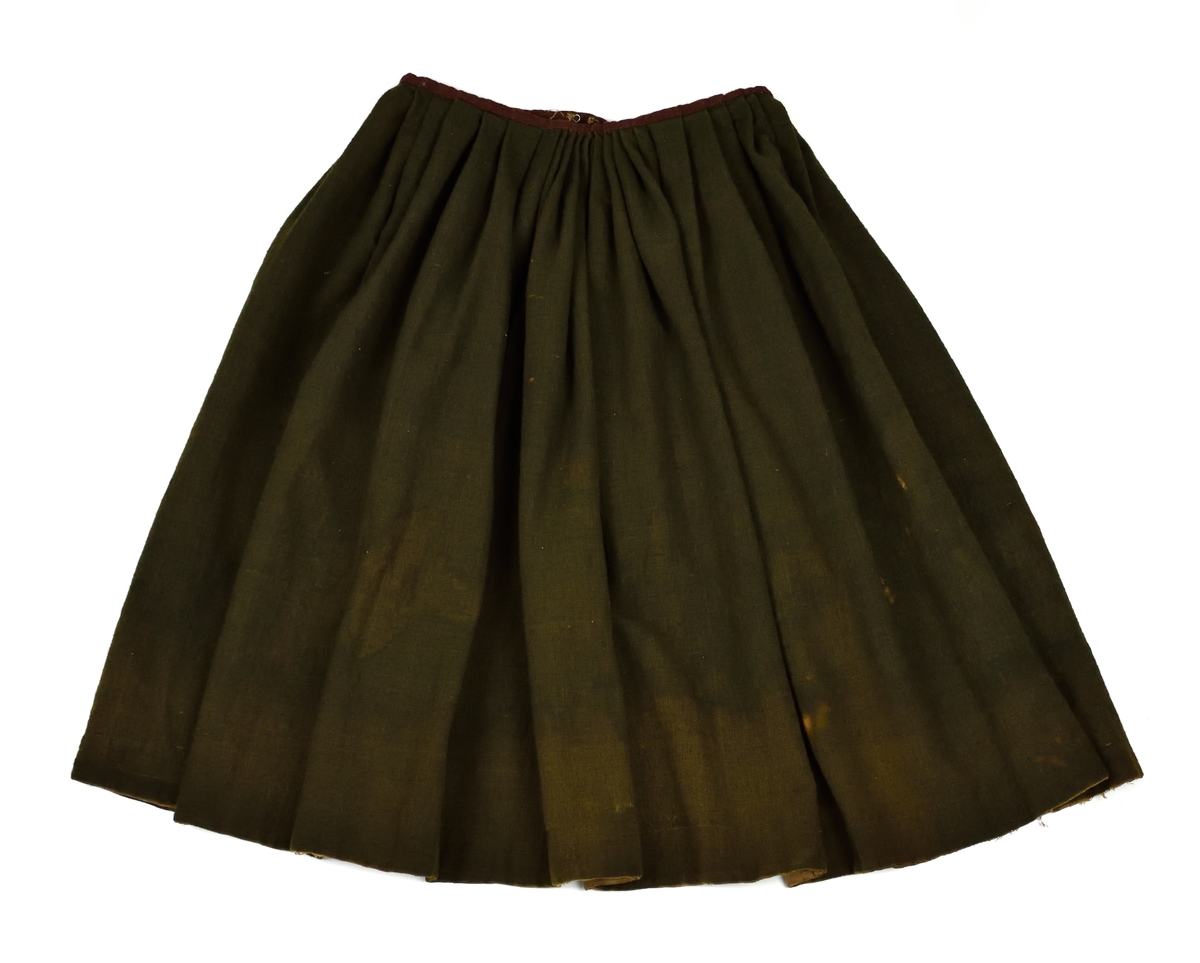 Handsydd kjol i svart halvylle med smal linning i rött ylle. Fållen är skodd med naturfärgat linne och en påsficka av grovt linne är infälld i en sidosöm.
