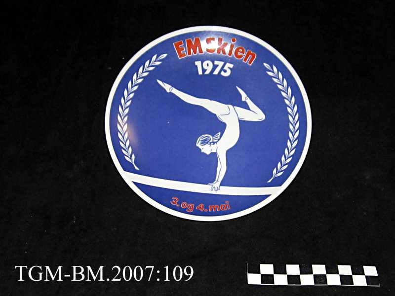 Platte i porselen, blå bunnfarge (glasur) med hvit turner (jente) og teksten: "EM Skien - 1975 - 3. og 4.mai"