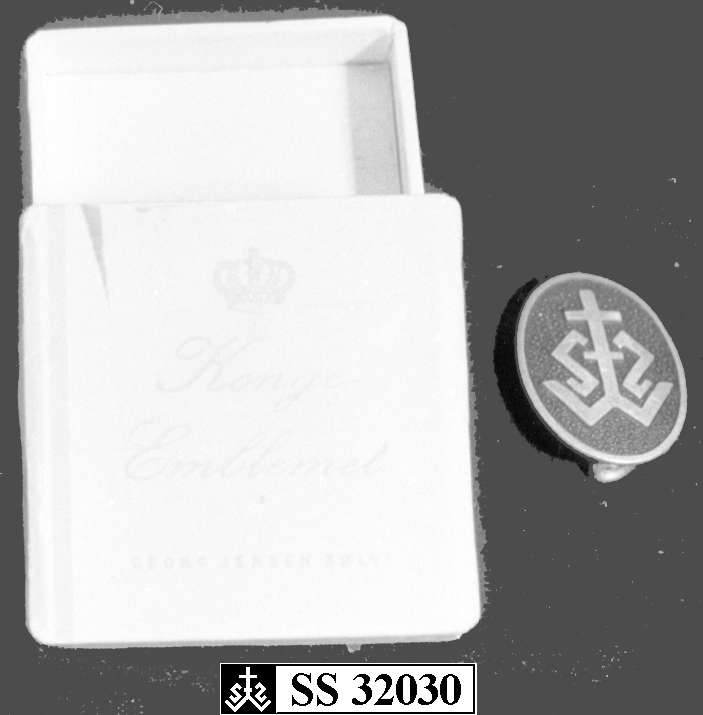 Jakkenål av sølv med blå bakgrunn og logo for De Sandvigske Samlinger. Nålen ligger i en eske som sansynligvis ikke er den originale emballasje for nålen.