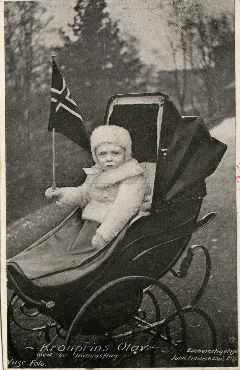 Postkort. Nyttårshilsen. Fotografisk sort/hvitt motiv. Kronprins Olav sitter i barnevogn med et norskt flagg. Stemplet 30.12.1905.