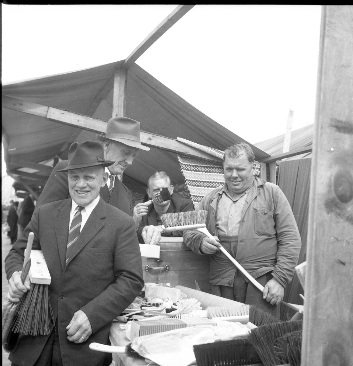 Marknad på Gränna torg. Borsthandlare Olle "Borsta-Olle" Bunnstrand säljer borstar av olika slag till kostymklädda herrar med slips och hatt. Till vänster står Erik Karlsson med borste i famnen och Axel Ahlin handlar en borste.
