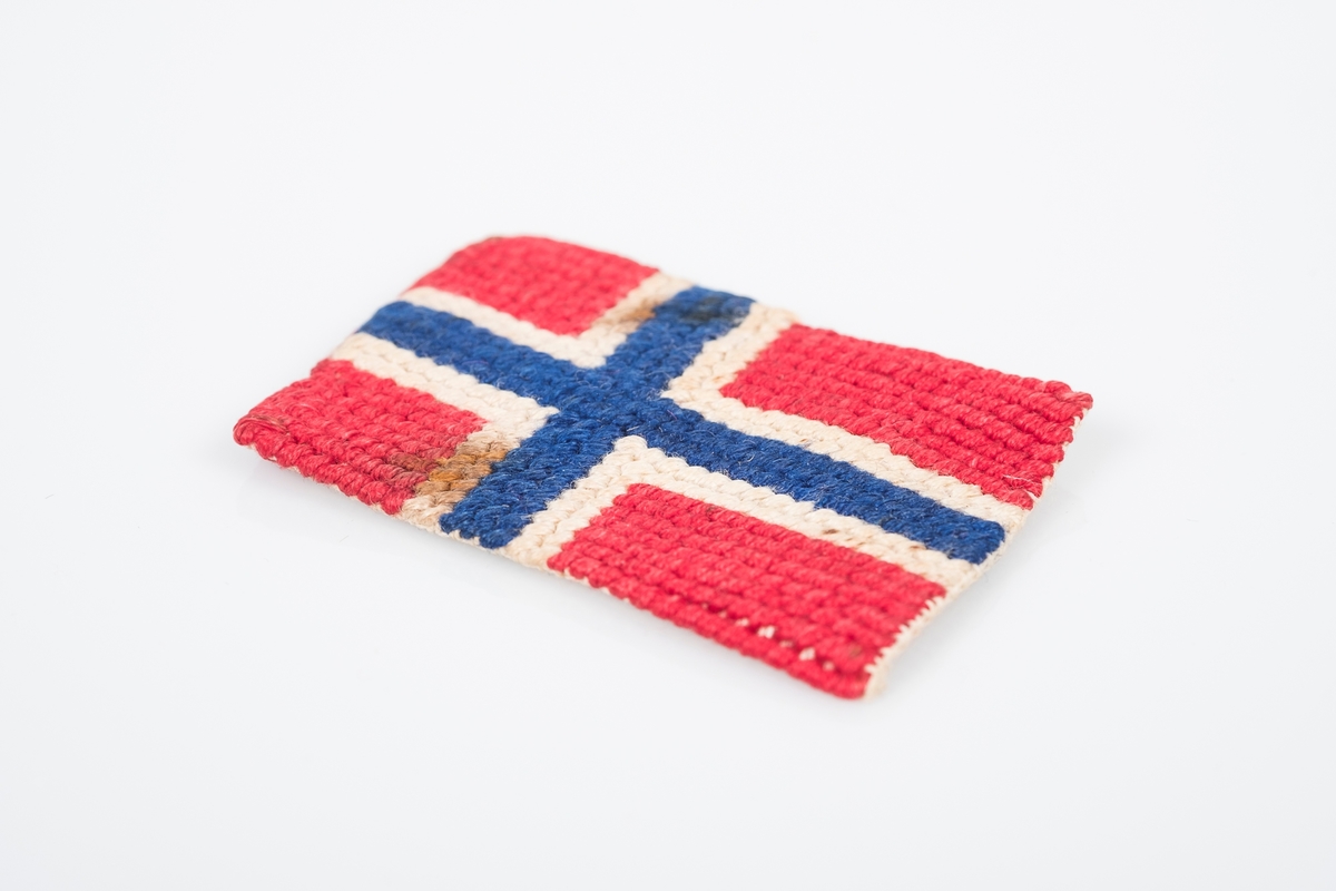 Norsk flagg brodert på en liten stoffbit, noen mørke flekker.