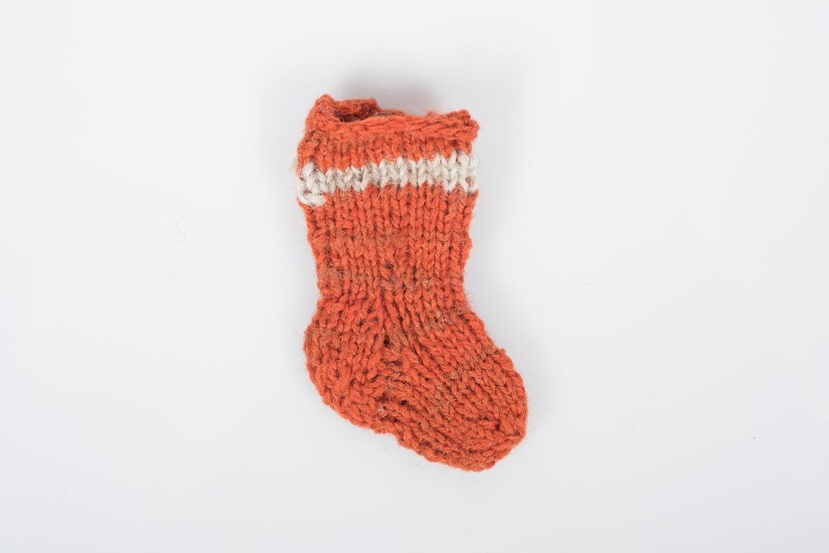 2. sokk av et par bittesmå strikkede ullsokker. Sokkene er oransjerøde med en lys stripe øverst og nøen brunrøde striper lenger ned.