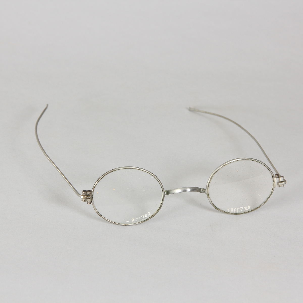 Glasögon, 1 par, med ovala glas och tunna vitmetallskalmar. Avlångt fodral av papp med rundade hörn, delbart.