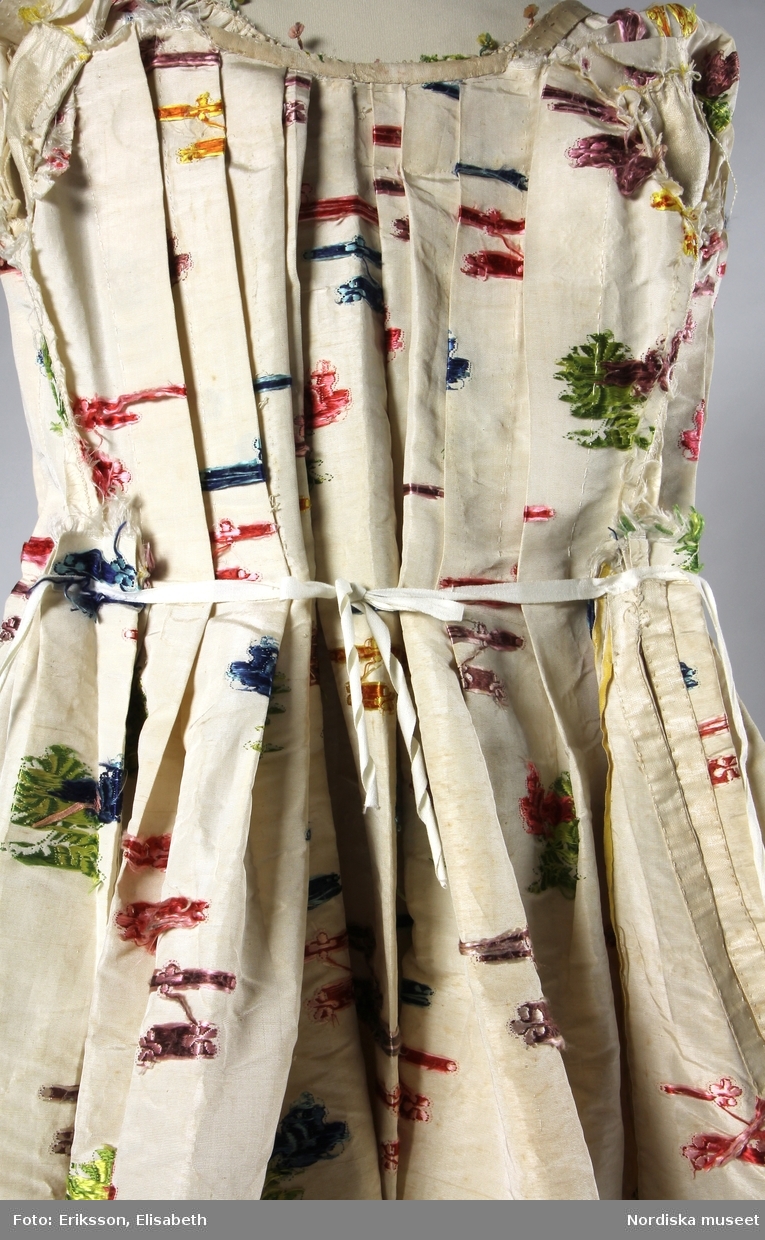 Brudklänning (robe), av broscherat, vitbottnat kinesiskt siden. Nygjord framvåd av fint vitt bomullstyg med kjolvolang nedtill av robens tyg. Två lösa garneringsbårder.