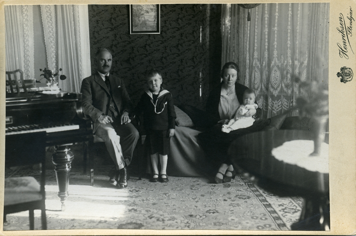 Familiebildet av kvinne og mann og to barn. De er avbildet sittende i en stue, med et flygel på den ene siden og lange hvite gardiner i bakgrunnen.