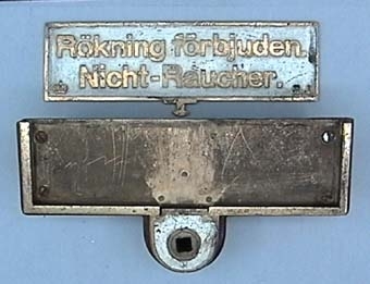 Vändbar tvåspråkig skylt i fastskruvad ram, med mässingstext i svenska och tyska på vit botten.
På ena sidan: "Rökare. Raucher."
På andra  sidan: "Rökning förbjuden. Nicht-Raucher."

Skylten sitter i en hållare av trä.